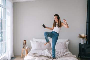 Active fit young woman in jeans  dancing on bed using headphones and cell phone at home on weekend. Pretty student girl enjoys music at hotel during vacations. Happy Italian female celebrates.