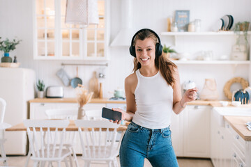 Cheerful hispanic young woman in casual using headphones and phone dancing at kitchen looks at camera toothy smiles happy by new home. Beautiful female student having fun at home.