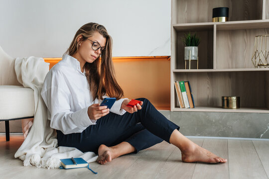 Pensive hispanic young adult woman in white shirt and dark blue pants sits on floor at living room holds phone and credit card makes order via internet against fireplace and book shelfs on background.