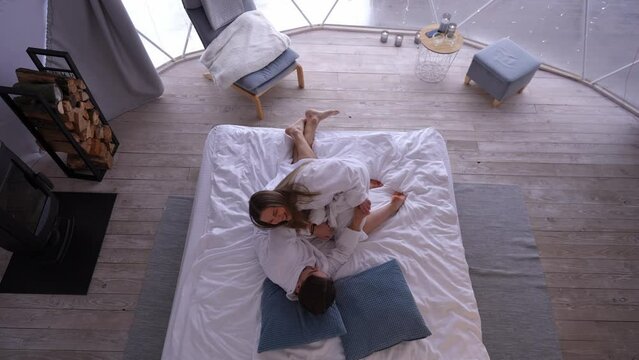 Top view joyful young Caucasian couple having fun cuddling on bed indoors. Wide shot carefree relaxed boyfriend and girlfriend in bathrobes laughing tickling in bedroom at home