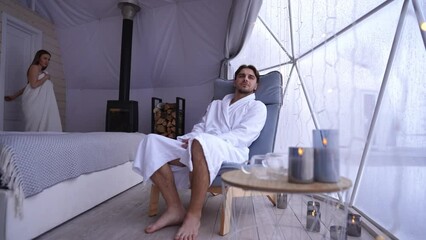 Relaxed man looking out the window sitting in bedroom as woman walking out bathroom in towel seducing. Caucasian young couple flirting indoors at home smiling
