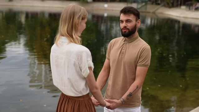 Tracking shot of young couple in love standing holding hands outdoors near beautiful lake at city park looking at each other smiling. Cheerful man and woman talking near water feeling happy.