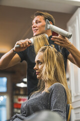 Fototapeta premium Hairstylist appointment. Young blond woman having her hair blown dried by professional hairdresser. Focus on the foreground. Vertical indoor portrait. Copy space. High quality photo