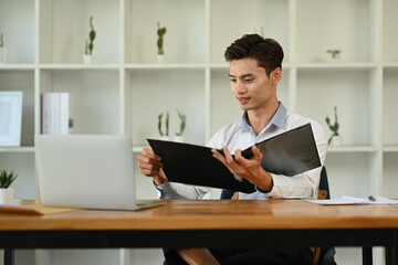 Concentrated asian man investor analyzing financial data and using laptop at his office desk