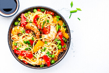 Stir fry noodles with shrimps, red and yellow paprika, green pea, chives and sesame seeds in ceramic bowl. Asian cuisine dish. White stone kitchen table background, top view