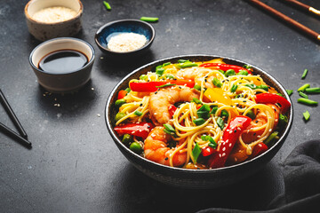 Stir fry noodles with shrimps, red and yellow paprika, green pea, chives and sesame seeds bowl. Asian cuisine dish. Black stone kitchen table background, top view