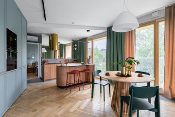 Creative composition of dinning room interior with kitchen, round table, green chairs, wooden kitchen island, red brastools, curtain, blue built kitchen and personal accessories. Home decor. Template.