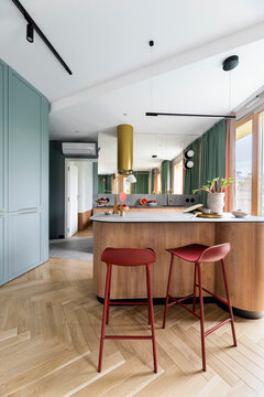 Modern composition of kitchen interior with wooden kitchen island, red barstools, colorful sculpture, green curtain, gold cooker hood, vase with rhubarb and personal accessories. Home decor. Template.