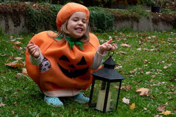 A laughing girl in a pumpkin costume with a lantern in her hand for Halloween on the background of nature.