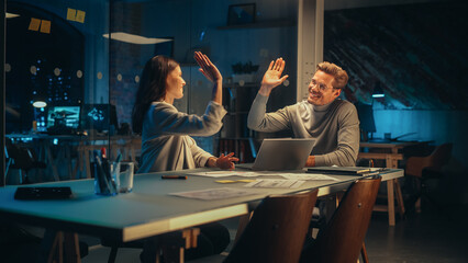 Successful Young Small Business Owners Communicating at a Meeting in Office at Night. Beautiful Female and Handsome Male Discussing Corporate Agenda and Opportunities. Managers High Five Each Other. - obrazy, fototapety, plakaty