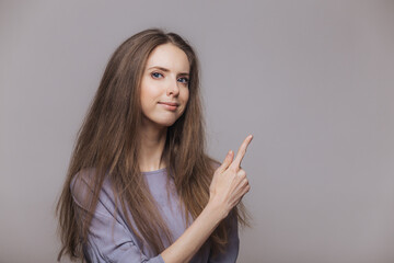 Shot of serious pleasant looking brunette female with blue eyes, dressed casually, points with index finger at blank copy space, isolated over grey background. Woman advertizes new product indoor