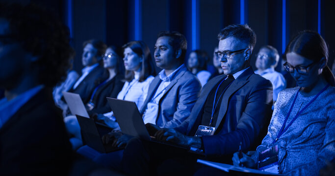 Young Man Sitting in a Crowded Audience at an IT Conference. Delegate Using Laptop Computer to Take Notes. Software Developer Watching Presentation About New High Tech Products and Cloud Solutions.