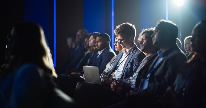 Male Sitting in a Dark Crowded Auditorium at a Tech Conference. Professional Using Laptop Computer. Specialist Watching Innovative Technology Presentation About New Hardware and High Tech Solutions.