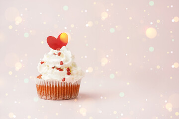 One homemade chocolate muffin with white creamy frosting and heart-shared red and white sugar decoration on marble board on pink surface  with bokeh and lights, space for text