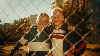 Portrait of Two Young Handsome Caucasian Boys Embracing Each Other while Looking at Camera, Smiling. Younger Brother Holding a Soccer Ball. Kids Standing Next to Eastern European Apartment Buildings.