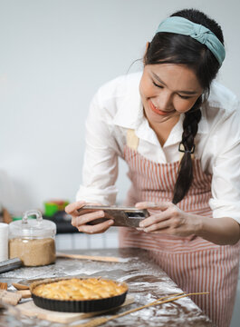 Young woman wears apron taking selfie photo with homemade pie in kitchen. Portrait of beautiful Asian female baking dessert and having fun taking photo by smartphone for online social. Home cookery.