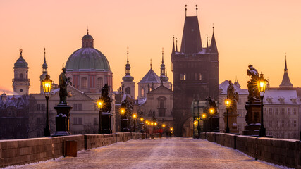 Charles Bridge covered in snow with shining lanterns in Prague during golden hour in the winter morning.