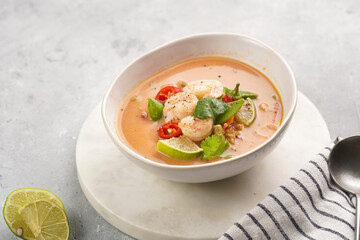 a spicy thai coconut soup or curry made of coconut milk, cherry tomatoes, snow pea, chili pepper, lime, ginger, shrimps and fresh cilantro on marble board on grey concrete background