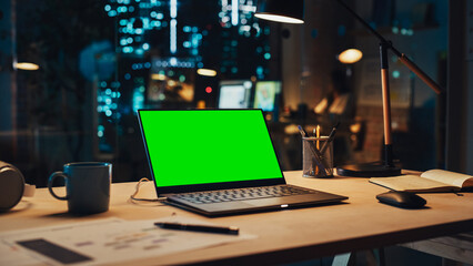 Laptop Computer Display with Mock Up Green Screen on a Table in Agency Office in the Evening. Chroma Key Monitor with Professional Working in the Background.