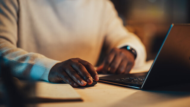 Close Up On Hands Of A Black Man Using A Mouse And Laptop In An Office At Night. Black African Specialist Browsing Internet, Researching Financial Data, Answering To A Work Email.