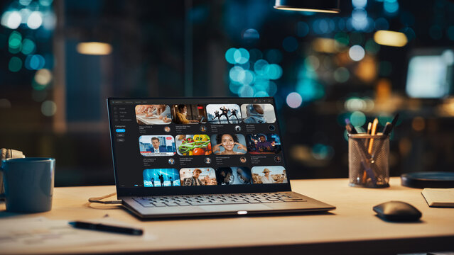 Laptop Standing on a Desk with a Video Streaming Platform Displayed on Screen. Table with Computer, Coffee Mug, Headphones and Notebook in Creative Office at Night.