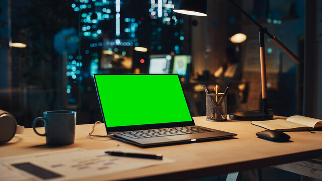 Laptop Computer Display with Mock Up Green Screen on a Table in Agency Office in the Evening. Chroma Key Monitor with Professional Working in the Background.