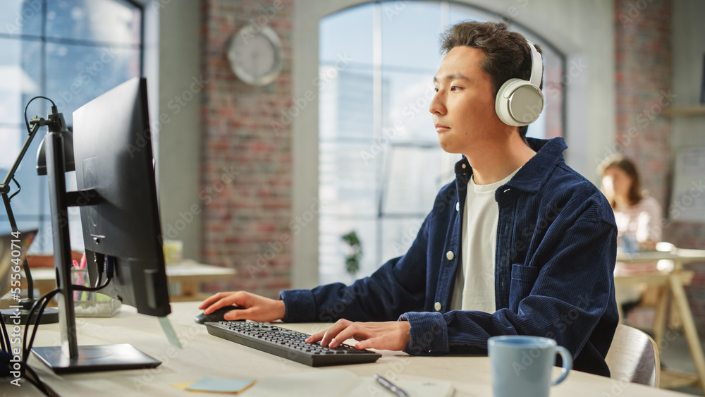 Fototapete Portrait of Asian Creative Man Using Computer and Headphones in Bright Modern Office. Male Data Analyst Developing Database While Listening to Music.