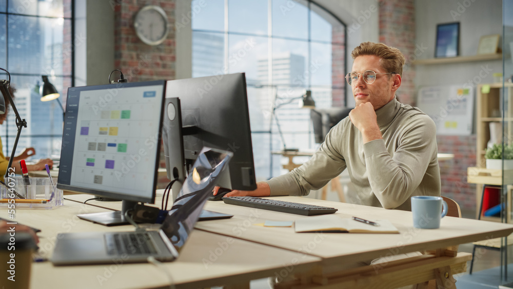 Poster Portrait of Focused Caucasian Young Man With Glasses Working on a Computer in Office. Male Director Preparing a Marketing Project. Diverse Team Working on Computers in the Background.