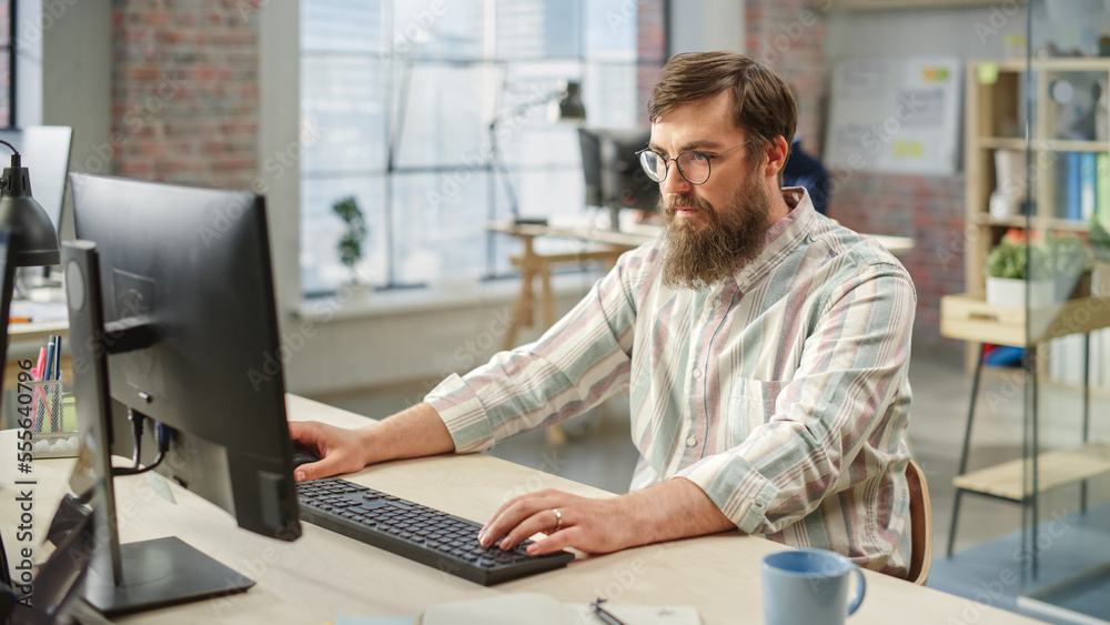 Poster portrait of young caucasian bearded man with glasses focused while sitting and working on computer i