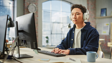 Portrait of Asian Male Employee Working on Computer in a Modern Office During the Day. Customer Support Agent Answering Requests While Using Headphones.