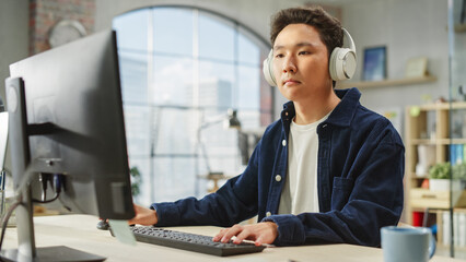 Portrait of Asian Creative Young man Focused While Using Computer in Modern Office. Logistics Supervisor with Headphones on, Analyzing Charts, Stock Market and Financial Data