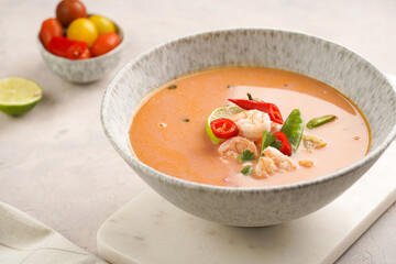 a spicy thai coconut soup or curry made of coconut milk, cherry tomatoes, snow pea, chili pepper, lime, ginger, shrimps and fresh cilantro on marble board on grey concrete background