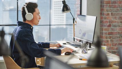Profile View of a Portrait of an Asian Young Man Working on a Computer and Using Headphones in a Modern Office. Data Analyst Reviewing Charts and Statistics.