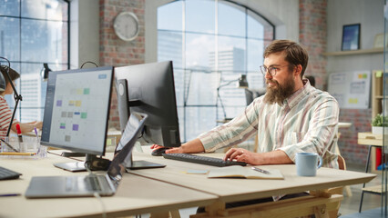 Portrait of Creative Bearded White Man with Glasses Smiling While Using Computer in Bright Spacious...