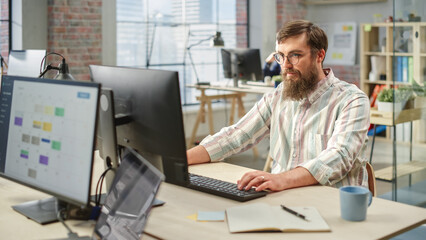 Portrait of Creative Bearded White Man with Glasses Focused While Using Computer in Bright Spacious Office. Marketing Manager Building a New Strategy.