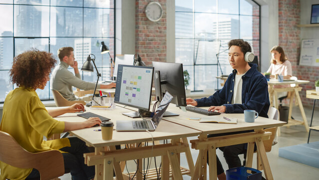 Multiethnic Creative Colleagues Working Using Computers in an Open Office During Day Time. Asian Man Listening to a Meeting on Headphones. Female Director working on a Schedule. - Powered by Adobe