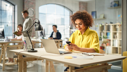 Portrait of Young Biracial Woman Working With a Laptop in a Bright Spacious Office. Female Production Lead Receiving an Important Work Notification on her Smartphone.
