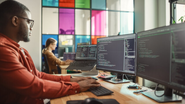 African American Man Writing Code on Desktop Computer With Multiple Monitors Set Up and a Laptop in Creative Office. Professional Data Scientist Using Software to Analyze Information From Internet. - Powered by Adobe