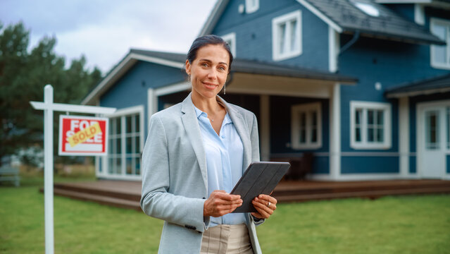 Portrait of an Intelligent Real Estate Agent in Smart Suit Holding Tablet Computer, Looking at Camera and Smiling. Multiethnic Realtor Standing in Front of Residential Property.