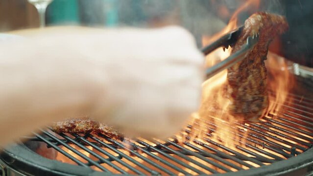 Person Using Tongs To Turn Over Juicy Sirloin Steak On Flaming Hot BBQ Grill With Smoke Rising From It. Low Angle, Static Shot