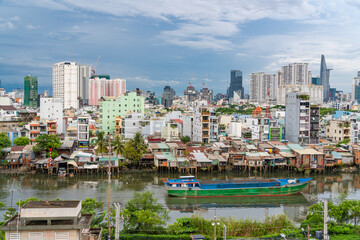 Naklejka premium HO CHI MINH, VIETNAM - December 3, 2022: Slum wooden house on the Saigon river bank, in front of modern buildings in ho chi minh city. View to district 1, see Bitexco tower, Landmark 81.