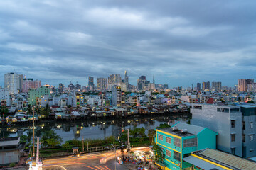 Fototapeta premium HO CHI MINH, VIETNAM - December 3, 2022: Slum wooden house on the Saigon river bank, in front of modern buildings at night in ho chi minh city. View to district 1, see Bitexco tower, Landmark 81.
