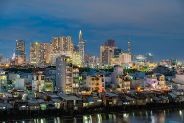 Plakat HO CHI MINH, VIETNAM - December 3, 2022: Slum wooden house on the Saigon river bank, in front of modern buildings at night in ho chi minh city. View to district 1, see Bitexco tower, Landmark 81.