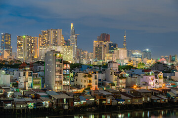 HO CHI MINH, VIETNAM - December 3, 2022: Slum wooden house on the Saigon river bank, in front of modern buildings at night in ho chi minh city. View to district 1, see Bitexco tower, Landmark 81.