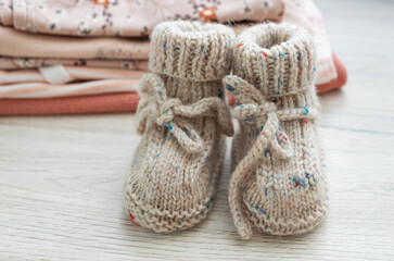 Knitted socks, knitted children's shoes made of natural wool, near a stack of children's clothes on a wooden background. Knitting, needlework, hobby.