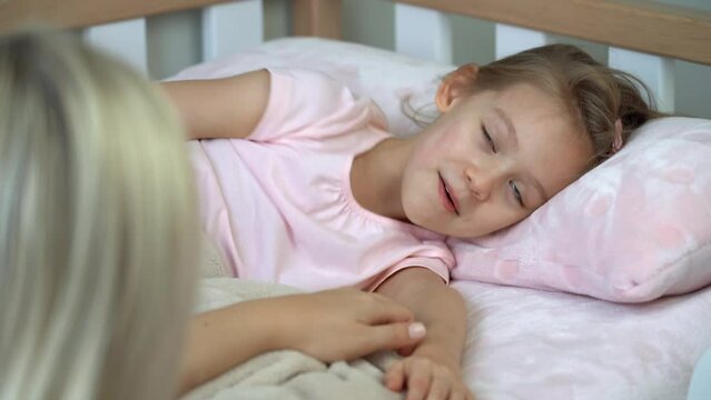 Back view of mother sits next to little daughter in bed and puts sleep. Child laughs and naughty before bed. Healthy sleep concept