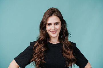 Fototapeta premium Beautiful brunette woman in black sweater looking at camera touches hair looking aside over studio turquoise background. Fashionable fit hispanic girl posing at studio. Fashion and beauty. Mockup