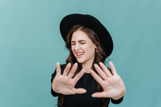 Beautiful brunette young woman in hat with brim stretches hands forward trying to stop someone, eyes closed with annoyed face expression over studio backdrop. Isolated hipster girl refusing.