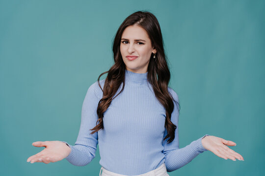 Unhappy brunette young woman with long wavy hair dressed in lilac sweater spreads hands in perplexity, upset stands over turquoise studio background. Peoples honest emotions. Mockup