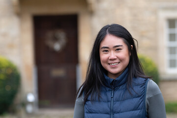 Fototapeta premium Smiling Asian woman stands in front of a traditional English house in the Cotswolds. Winter holiday portrait in the UK.